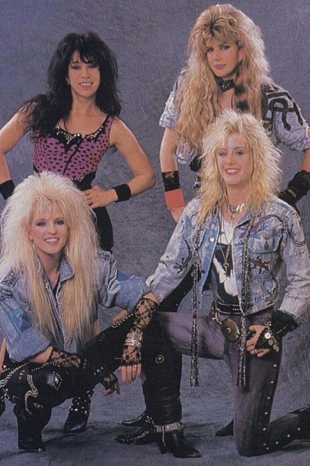 Four women with big, teased hair and 1980s rock fashion pose together. They wear denim, studded accessories, and bold makeup, standing and kneeling in front of a gray backdrop, smiling at the camera.
