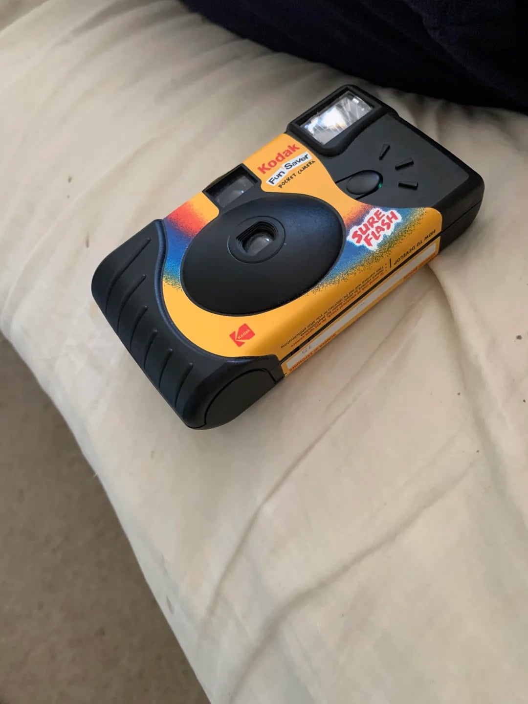 A Kodak disposable film camera with a yellow and black design labeled "Super Flash" rests on a light-colored bedsheet.