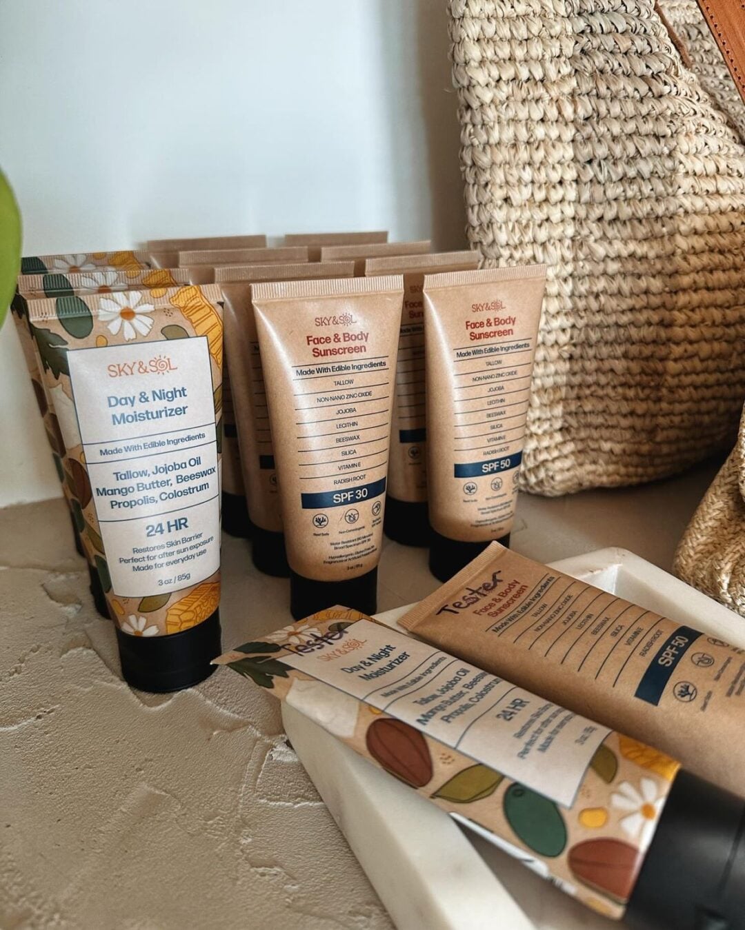 Several tubes of SKY & SOL skincare products, including moisturizers and sunscreens, are arranged on a light surface next to a textured woven bag. The tubes have natural-themed designs and labels.