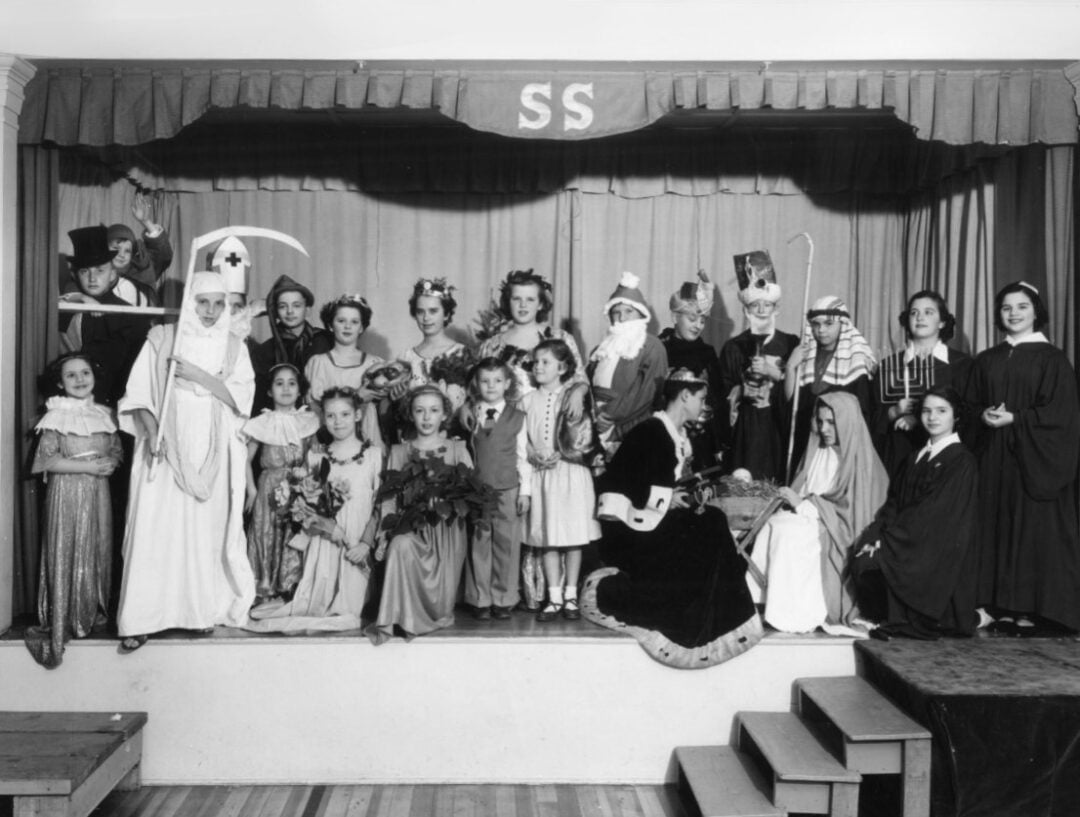 A group of children in costumes pose on a small stage with a curtain and a sign reading "SS" above. They are dressed as various characters, including a nurse, royalty, and biblical figures, suggesting a school play or pageant.