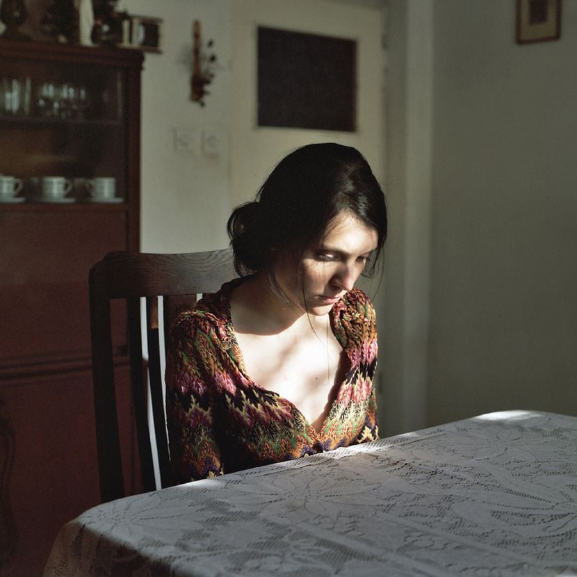 A woman with dark hair sits alone at a table covered with a lace tablecloth, looking down thoughtfully. Soft light from a window illuminates her face and shoulder, casting gentle shadows.