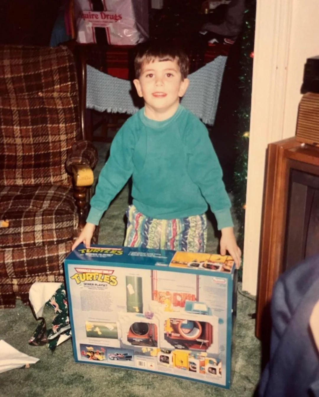 A young boy in patterned pajamas and a green shirt smiles while holding a Teenage Mutant Ninja Turtles toy box in a cozy living room with plaid furniture and holiday decorations.
