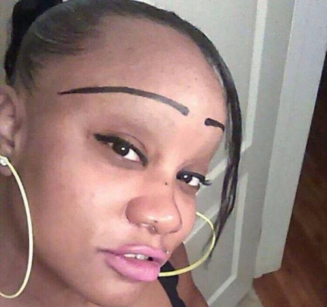 A woman with large yellow hoop earrings and pink lipstick poses for a selfie. Her eyebrows are drawn unusually high and straight above her natural brow line.