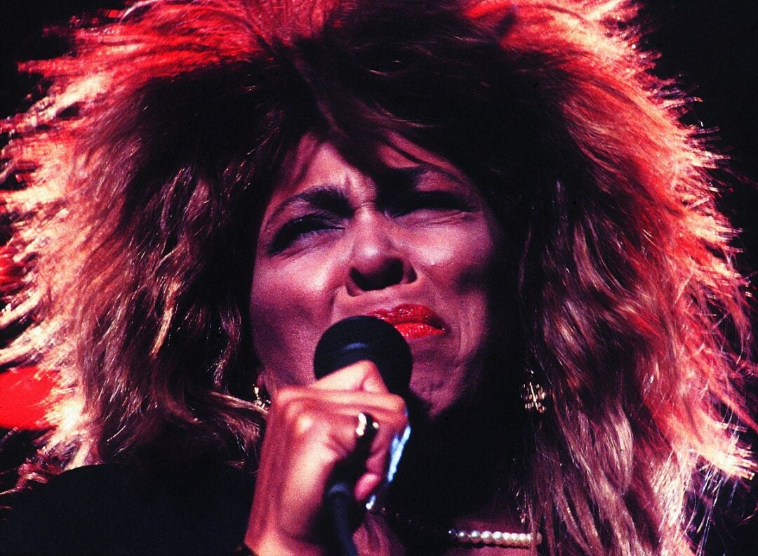 A singer with voluminous hair and red lipstick passionately performs with eyes closed, holding a microphone close to their mouth against a dark, brightly lit background.