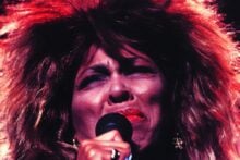 A person with big, wild hair and bright red lipstick sings passionately into a microphone on stage, eyes closed in emotion, with dramatic lighting highlighting their face.