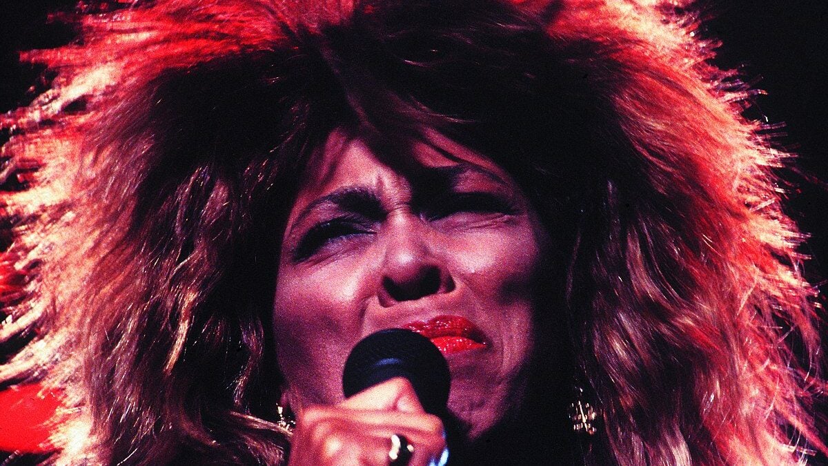 A person with big, wild hair and bright red lipstick sings passionately into a microphone on stage, eyes closed in emotion, with dramatic lighting highlighting their face.
