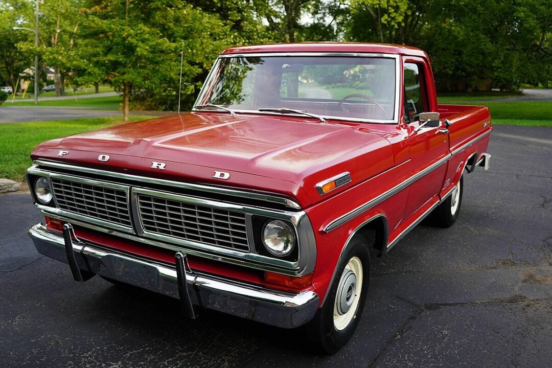 A vintage red Ford pickup truck is parked on a driveway, with green trees and grass visible in the background on a sunny day.