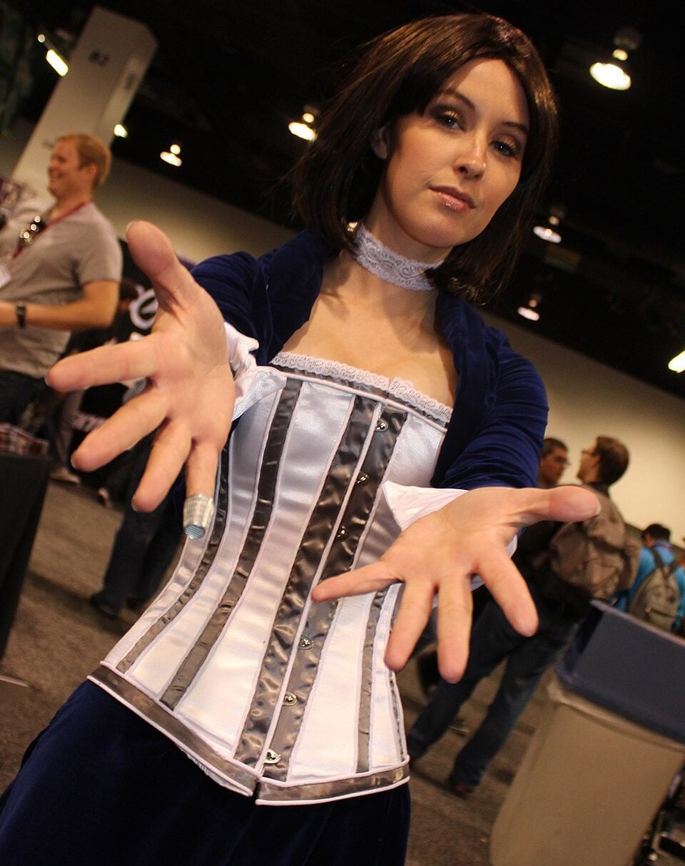 A woman in a blue and white cosplay costume extends her hands toward the camera at a convention, with other people and booths visible in the background.