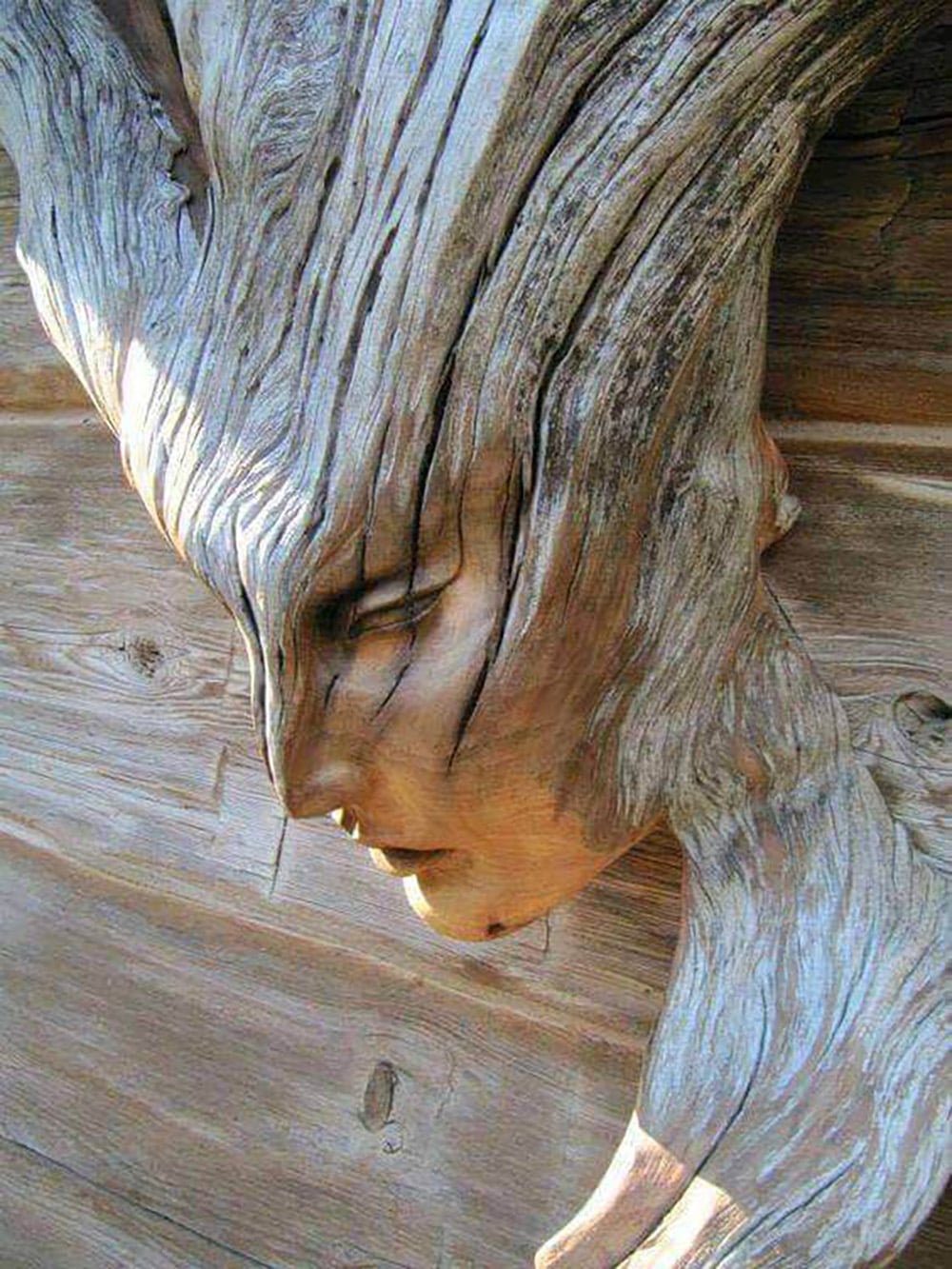 A close-up of a weathered wooden sculpture carved to resemble a human face, with flowing hair-like patterns blending into the wood grain on a wooden background.