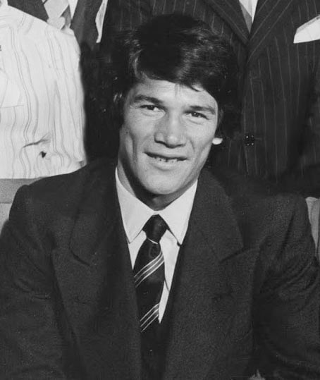 A man with dark hair wearing a suit and striped tie sits and smiles slightly, looking at the camera. Other people in suits are partially visible in the background.