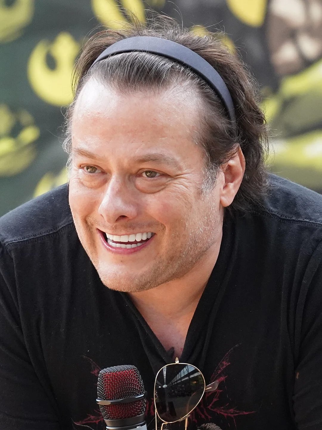 A man with light skin and brown hair, wearing a dark headband and a black shirt, smiles while speaking into a microphone at an outdoor event. Sunglasses hang from his shirt collar. The background is blurred with green and yellow patterns.
