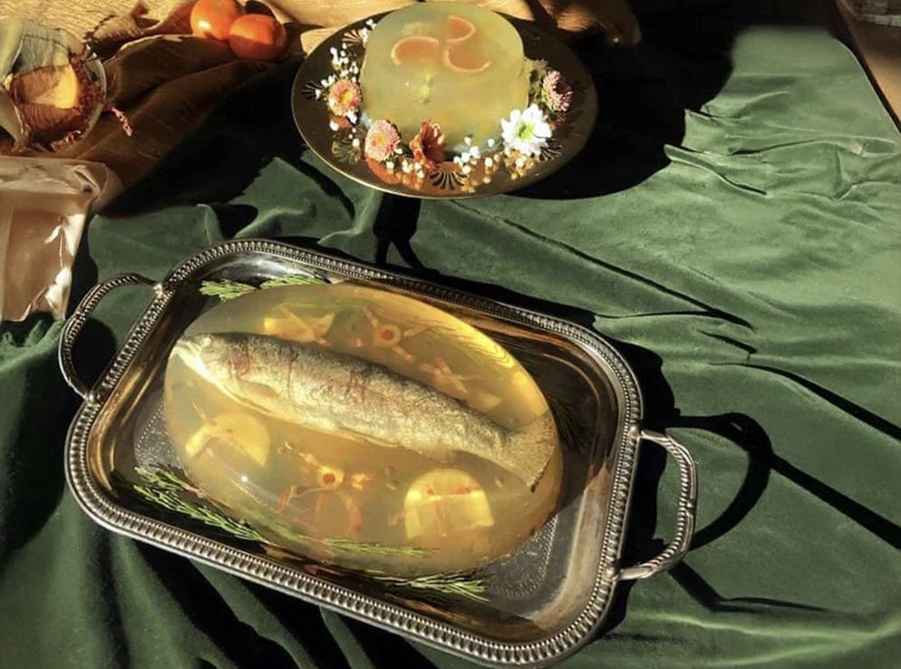 A whole fish suspended in clear aspic on a silver tray sits on green velvet; behind it, a decorated gelatin mold with mushrooms and herbs is displayed on a black plate.