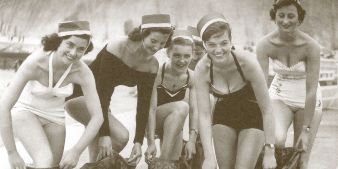 Five women in vintage swimsuits and hats smile and crouch in a playful pose on a beach, participating in an activity together. The image is in black and white, suggesting it is from a past era.