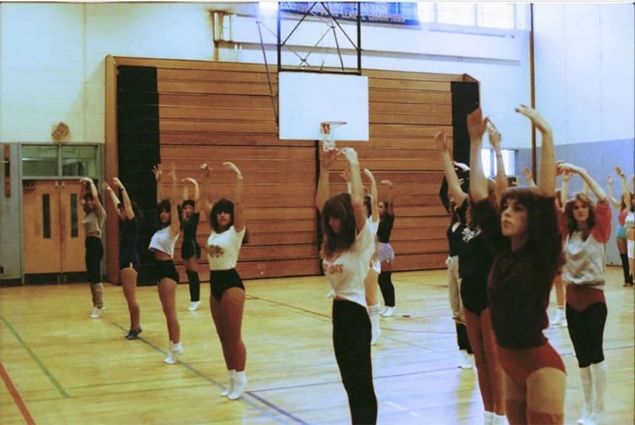 A group of people participate in an aerobics class in a gymnasium, stretching with arms raised. They wear leotards, shorts, and athletic wear, and a basketball hoop is visible in the background.