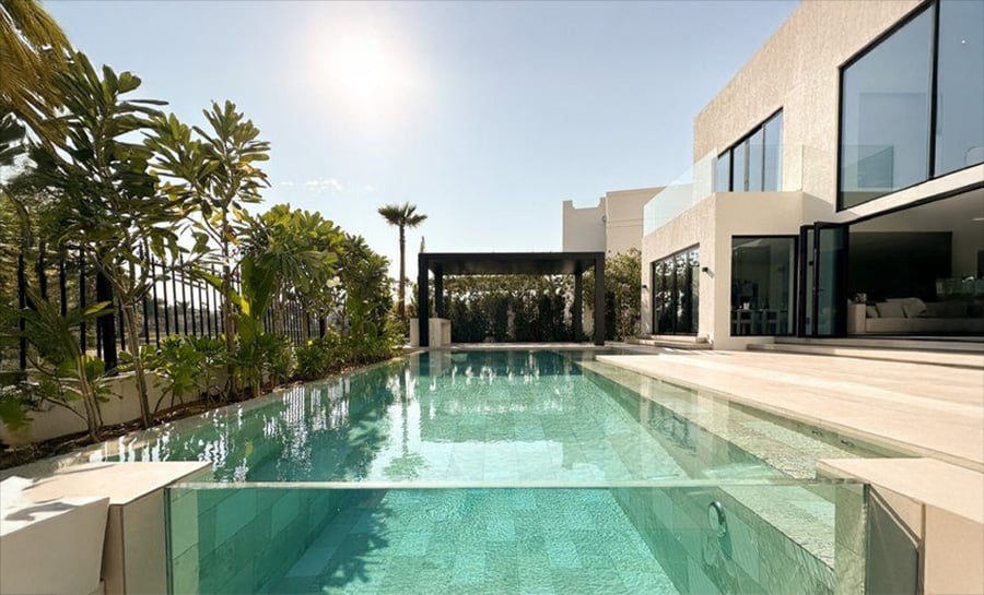 Modern luxury house with a large, clear swimming pool, surrounded by tropical plants and trees. Glass walls and open doors connect the indoor and outdoor spaces under a bright, sunny sky.