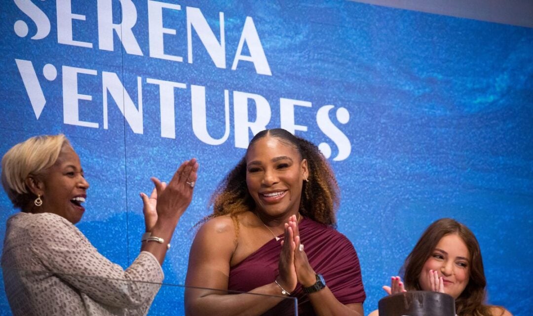 Three women stand smiling and applauding at a podium in front of a blue wall with "SERENA VENTURES" written on it. The podium has a sign reading "NEW YORK". One woman is clapping, another is smiling, and the third is looking on.