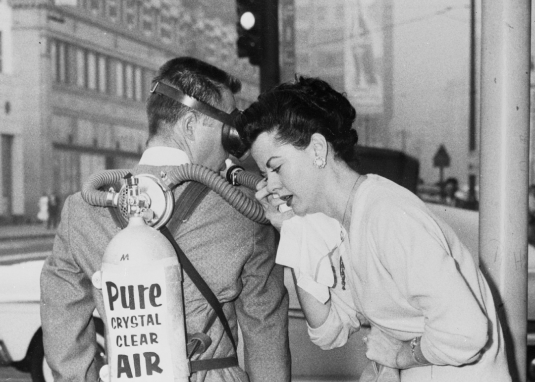 A woman covers her mouth with a handkerchief and leans toward a man wearing a suit and a tank labeled "Pure Crystal Clear Air" with a breathing mask, on a city street.