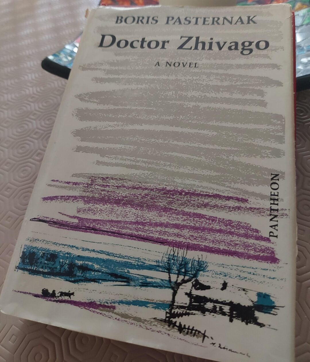 A copy of Boris Pasternak’s novel "Doctor Zhivago" lies on a table with a patterned tablecloth. In the background, there is a cups and a dish with colorful designs.