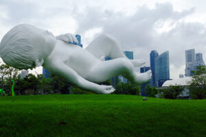 A large white sculpture of a floating baby lies on green grass with city skyscrapers and cloudy sky in the background. Trees and colorful structures are also visible on the left.