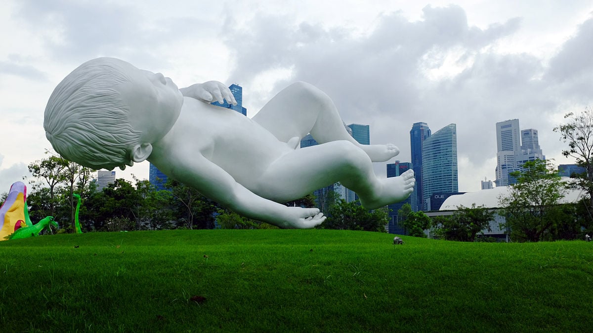 A large white sculpture of a floating baby lies on green grass with city skyscrapers and cloudy sky in the background. Trees and colorful structures are also visible on the left.