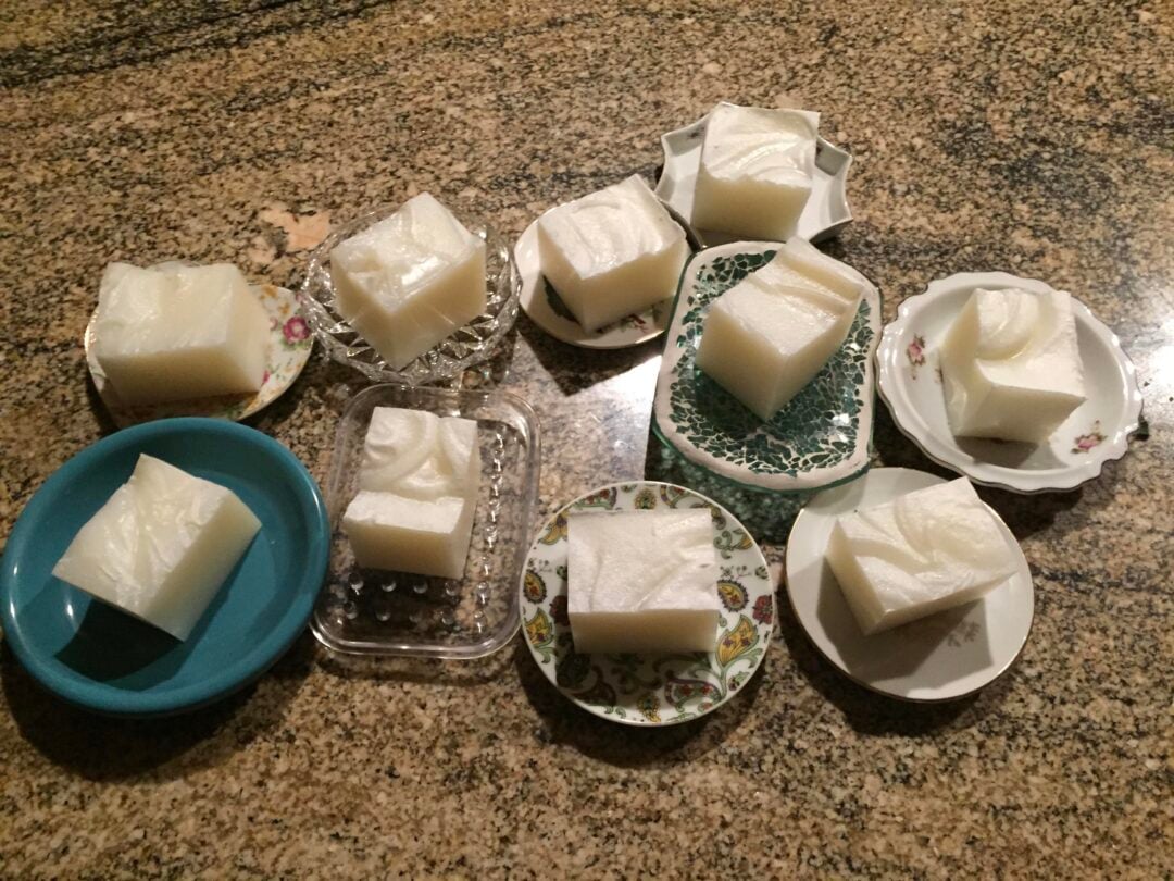 Nine square pieces of white gelatinous dessert, each served on a different small plate or dish, are arranged on a speckled brown countertop.