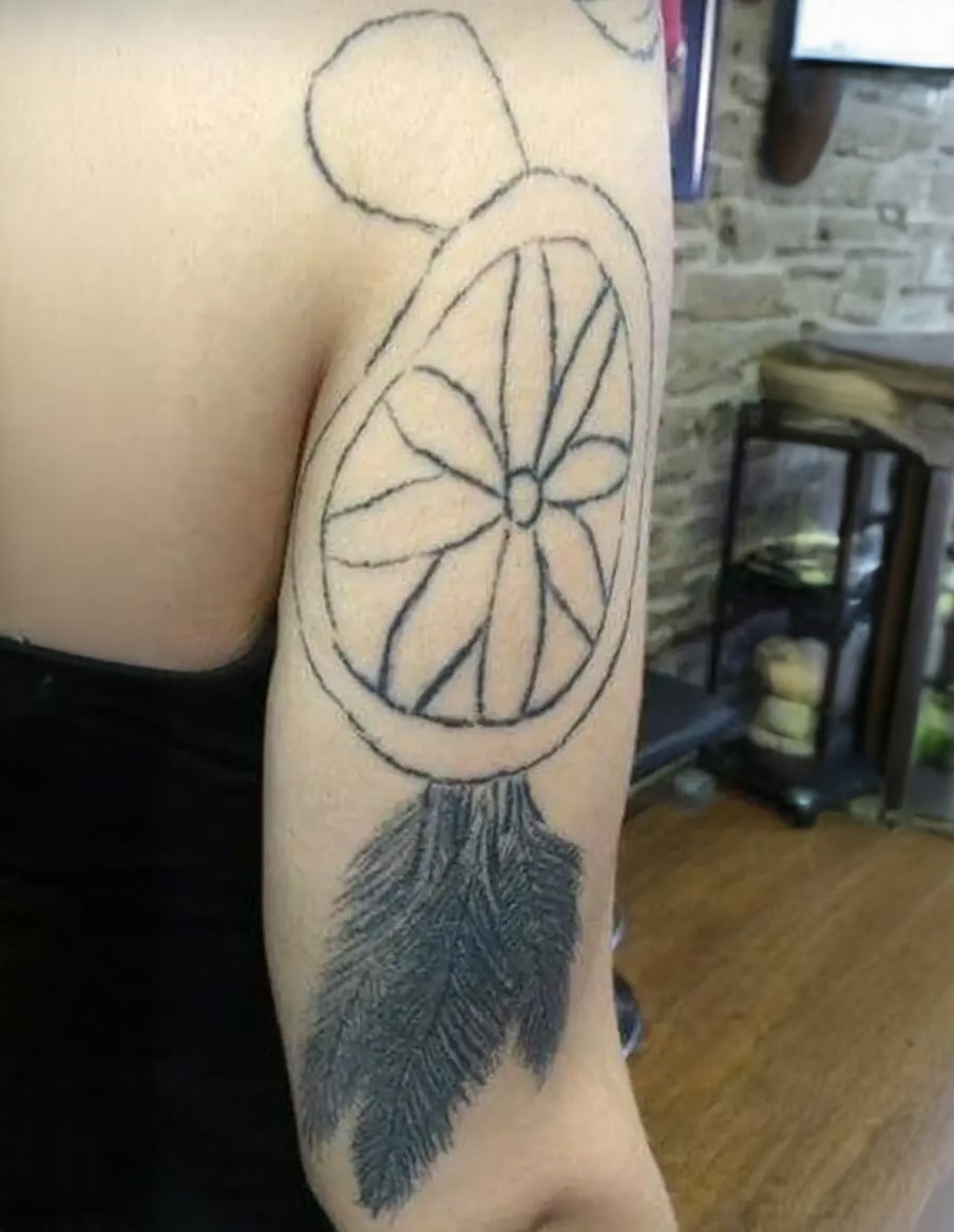 A person's upper arm with an unfinished tattoo outline of a dreamcatcher featuring a circular web design and detailed feather below, set in a room with exposed brick walls.
