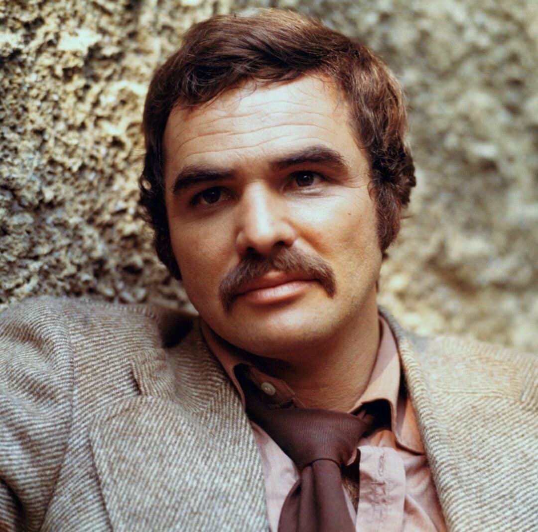 A man with brown hair and a mustache, wearing a light brown jacket, tan shirt, and loosened brown tie, sits against a textured stone wall and looks slightly off camera with a calm expression.