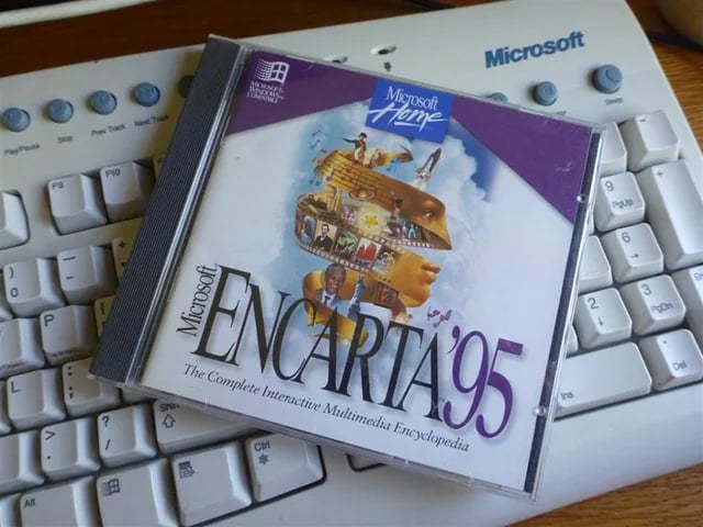 A CD case for Microsoft Encarta ’95, labeled “The Complete Interactive Multimedia Encyclopedia,” rests on a white Microsoft computer keyboard. The case features colorful graphics and the Microsoft Home logo.