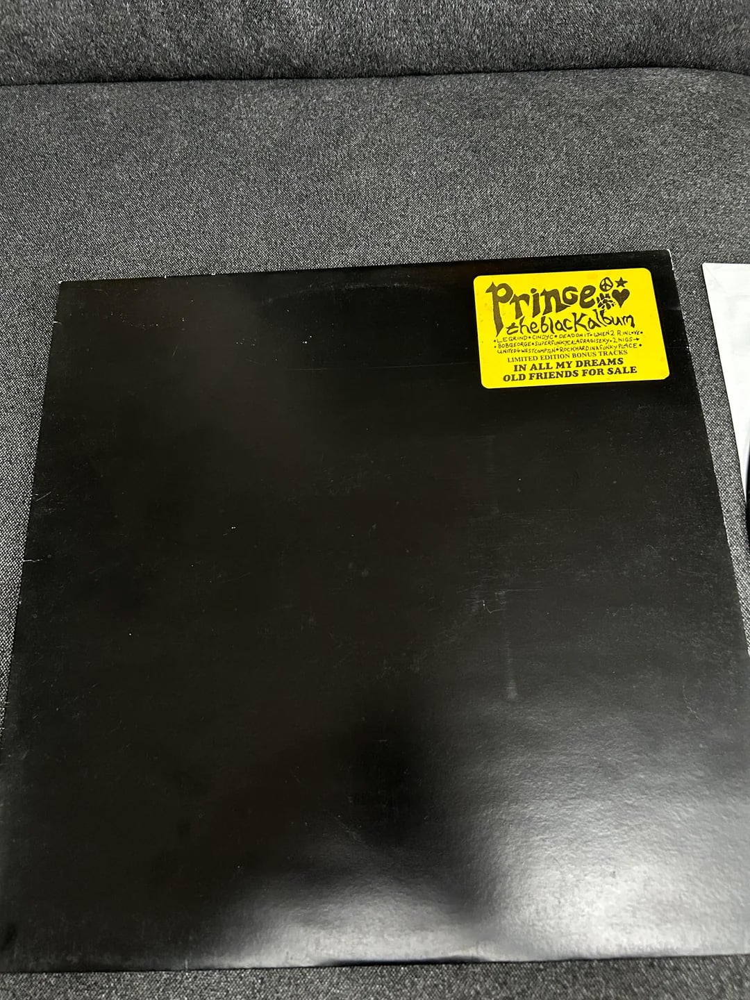 A black vinyl record sleeve with a yellow sticker reading "Prince The Black Album" in bold text, placed on a dark fabric surface. The sleeve is plain with no other visible text or images.
