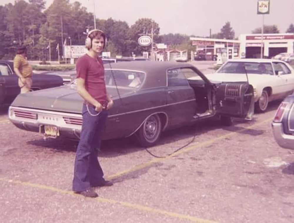 A man in a red shirt and jeans stands in a parking lot, wearing large headphones connected by a long wire to a green vintage car with its door open. Several other cars and a few people are visible in the background.