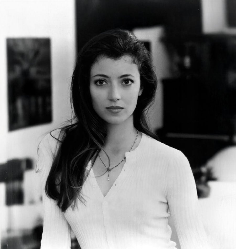 A young woman with long dark hair and a calm expression wears a light-colored buttoned top and a necklace, standing indoors with a blurred background. The photo is in black and white.