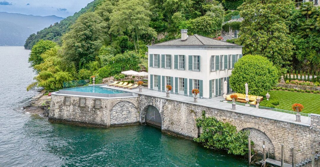 A white villa with blue shutters sits beside a lake, featuring an infinity pool, sun loungers, lush gardens, and stone walls, surrounded by trees and greenery on a hillside.
