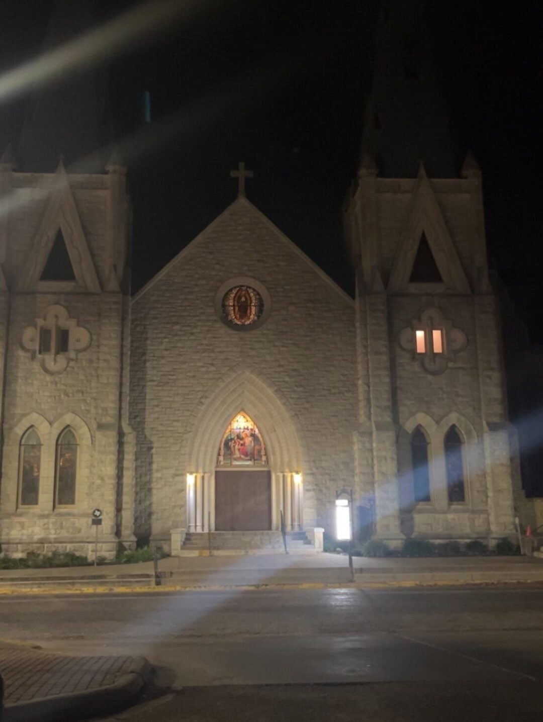 A large stone church with two tall steeples is illuminated at night. Warm light glows from the entrance and a few windows. The street and sidewalk in front are wet, reflecting the building’s lights.