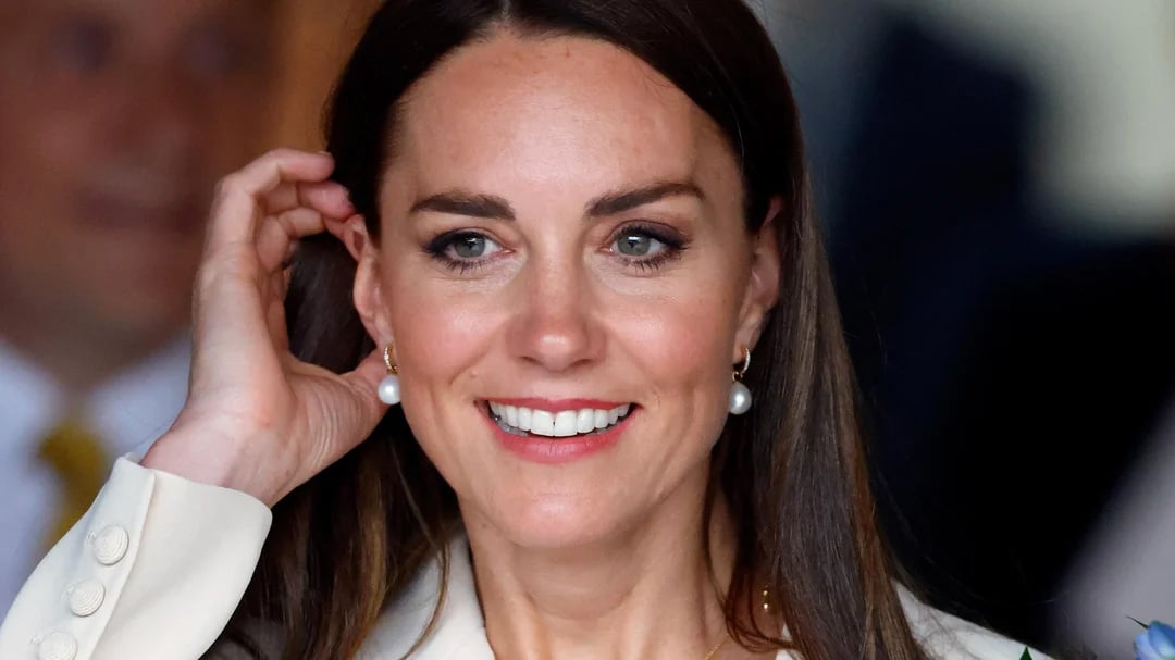 A woman with long brown hair and pearl earrings smiles brightly, looking slightly to the side while touching her ear. She wears a cream-colored outfit and is photographed in close-up with a blurred background.