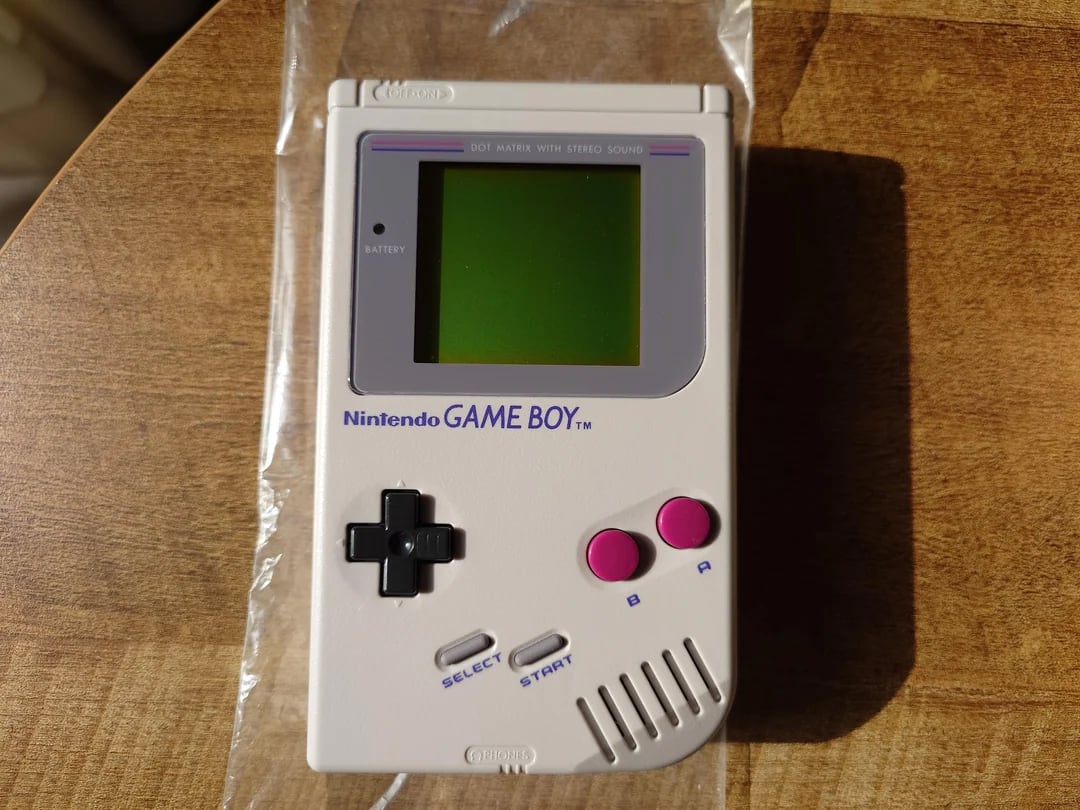 A classic Nintendo Game Boy handheld console with a gray body, black control pad, and pink A and B buttons, resting on a wooden surface in bright sunlight.