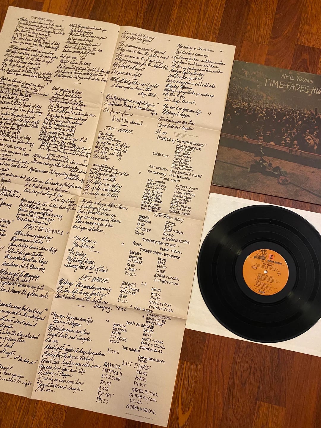 A vinyl record of Neil Young's "Time Fades Away" sits beside its album cover and an unfolded insert with handwritten lyrics and printed credits, all placed on a wooden floor.