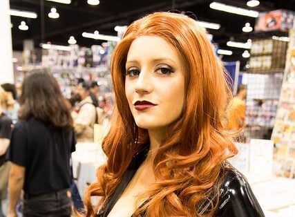 A woman with long red hair poses in a shiny black catsuit and gold accessories at a convention, with booths and other attendees visible in the background.