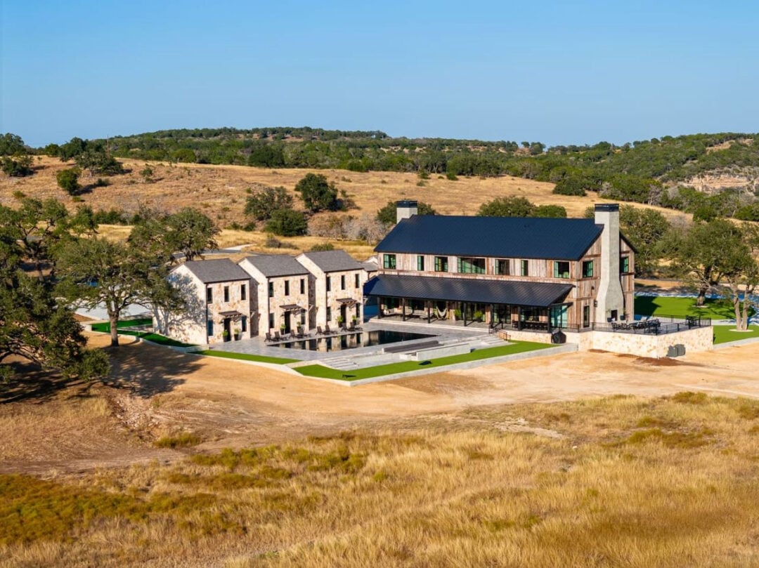 A large modern farmhouse with stone walls and black roof sits on open, grassy land with gently rolling hills and scattered trees under a clear blue sky. A separate building and swimming pool are nearby.