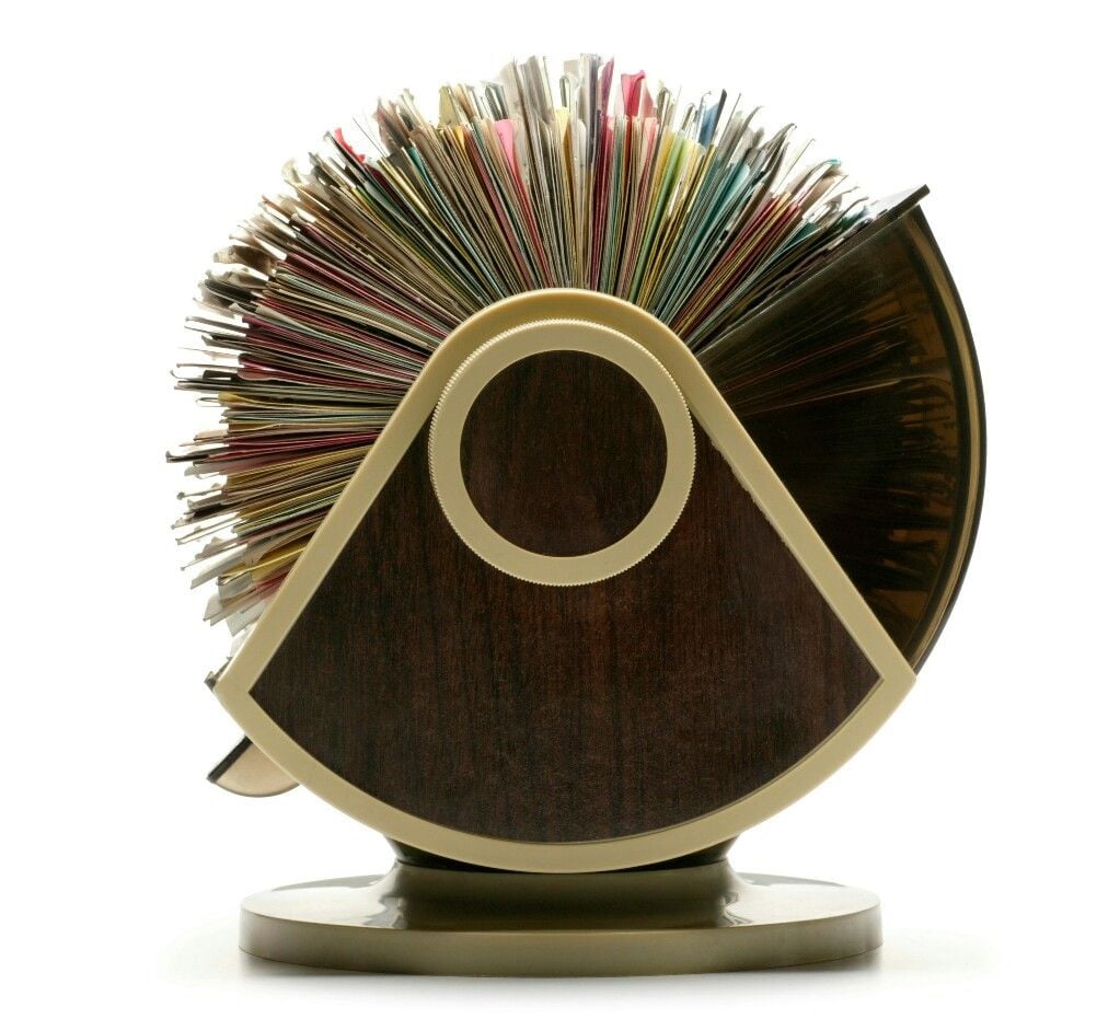 A vintage rotary card file with a circular design, filled with colorful index cards, stands on a brown and gold base against a white background.