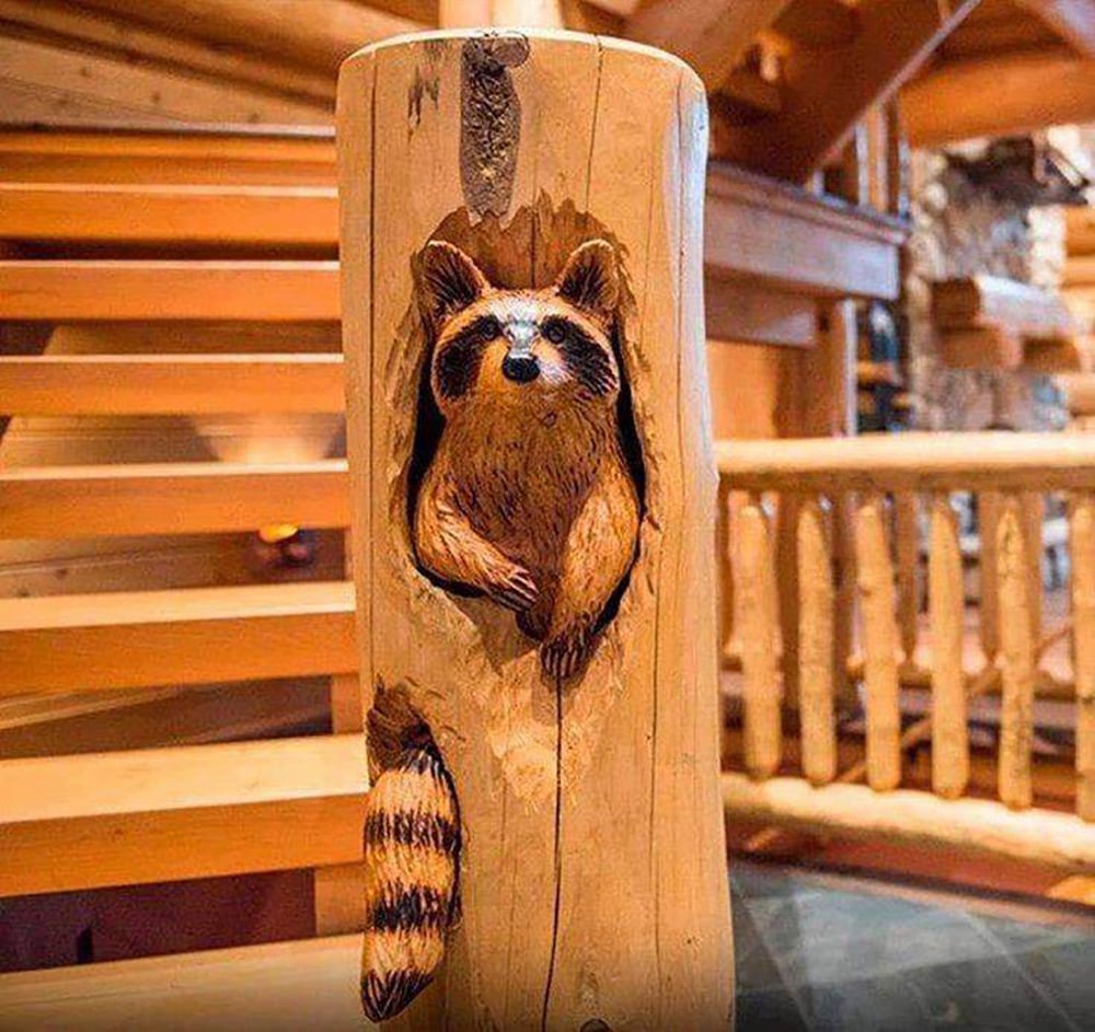 A wooden post in a log cabin features a detailed carving of a raccoon peeking out from a hollow, with its head, front paws, and striped tail visible. Stairs and wooden railings are in the background.