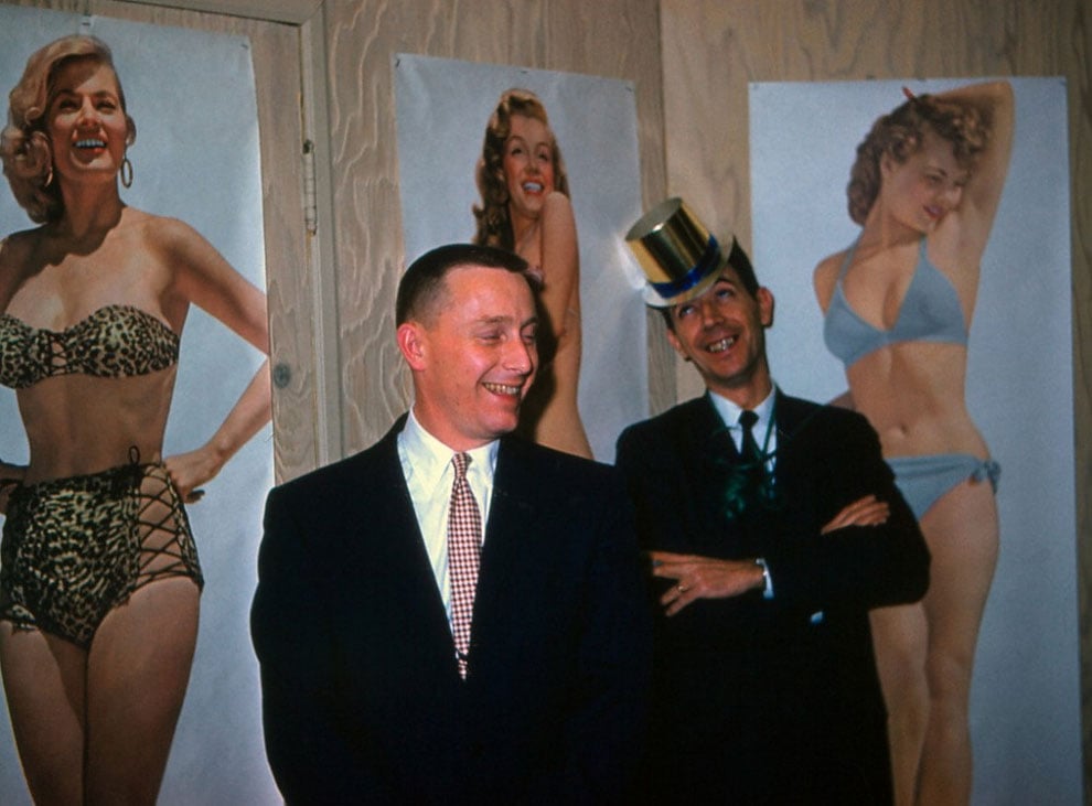 Two men in suits stand smiling in front of posters of women in swimsuits. One man wears a party hat and leans playfully, while the other stands upright and grins. The background features wood-paneled walls.
