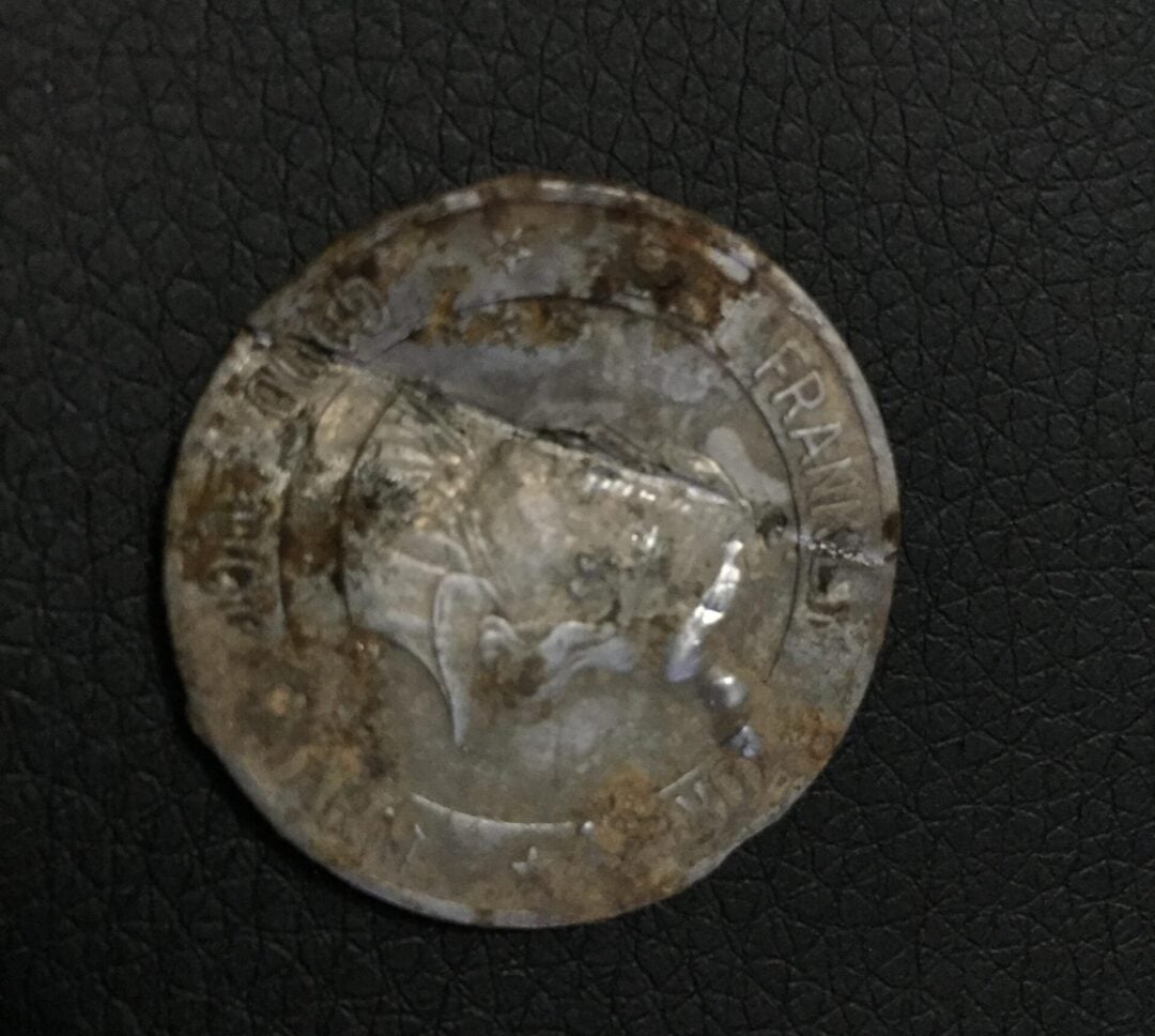 A worn, tarnished coin with a portrait in profile and partially readable text, resting on a textured black surface. The coin appears old and corroded, with visible scratches and discoloration.