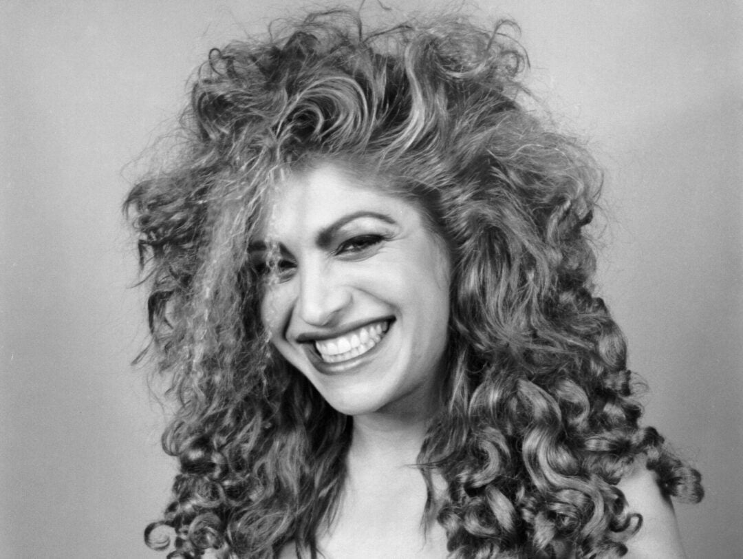 A woman with voluminous, curly hair smiles widely at the camera. She wears an off-the-shoulder black top and has a confident, cheerful expression. The photo is in black and white.