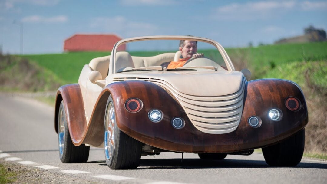 A person drives a unique convertible car with a wooden exterior design on a rural road, with green fields and a red building visible in the background.
