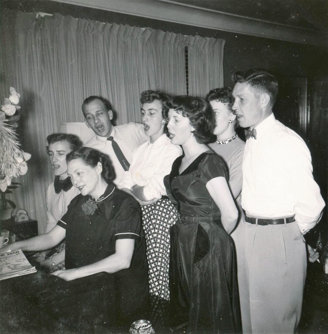 Six people stand around a piano, singing together while one woman plays. They appear to be enjoying themselves in a cozy, mid-century living room, dressed in 1950s-style clothing. Curtains and a floral arrangement are visible.