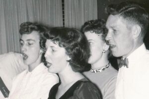 Six people stand and sing around a seated woman playing piano in a living room. Everyone appears engaged and joyful. The photo is black and white, with a curtain and chandelier visible in the background.