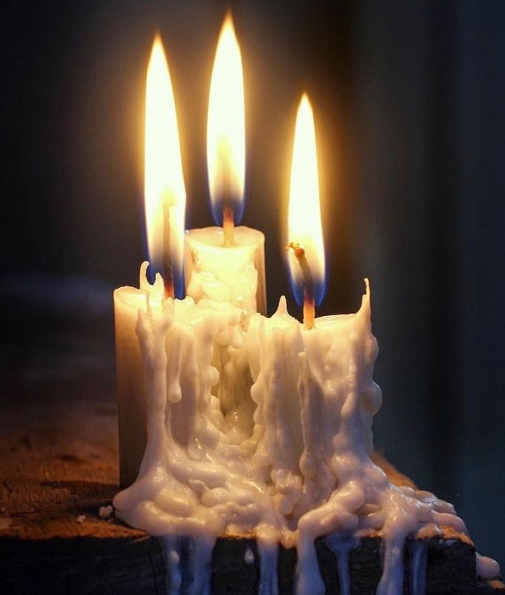 Three white candles burning side by side, with melted wax dripping down and pooling heavily, creating a textured, layered appearance. The background is dark, highlighting the candle flames.