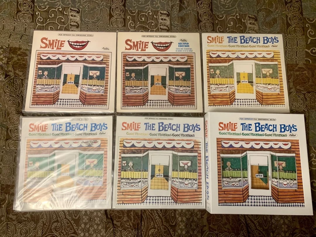 Six copies of The Beach Boys' "Smile" box set are arranged in two rows on a patterned bedspread. Each box features colorful artwork of a storefront with cartoonish design and smiling mouth logo.