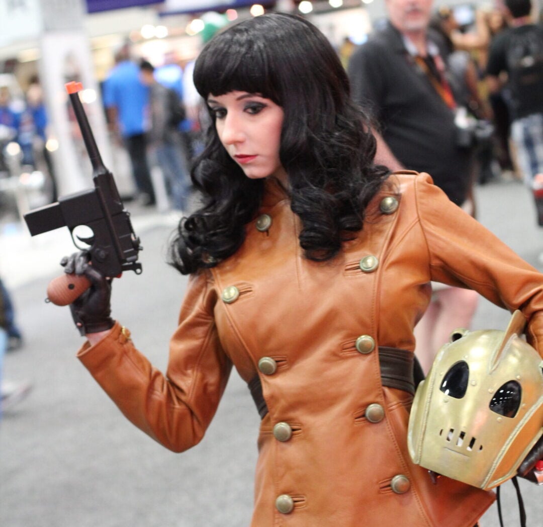 A person in a brown leather jacket, black wig, and stockings poses with a prop gun and a gold helmet at a convention, cosplaying as a character from "The Rocketeer.