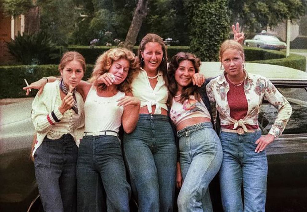 Five young women in 1970s-style clothing stand in front of a car, smiling and posing playfully together outdoors, with greenery and trees in the background.